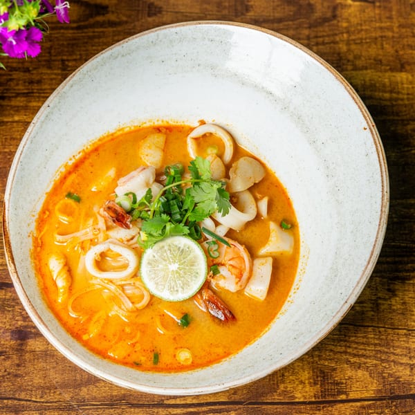 Jaew Thai Cuisine's Tom Yum Goong with shrimp, squid, scallops, noodles, and lime in a white bowl.